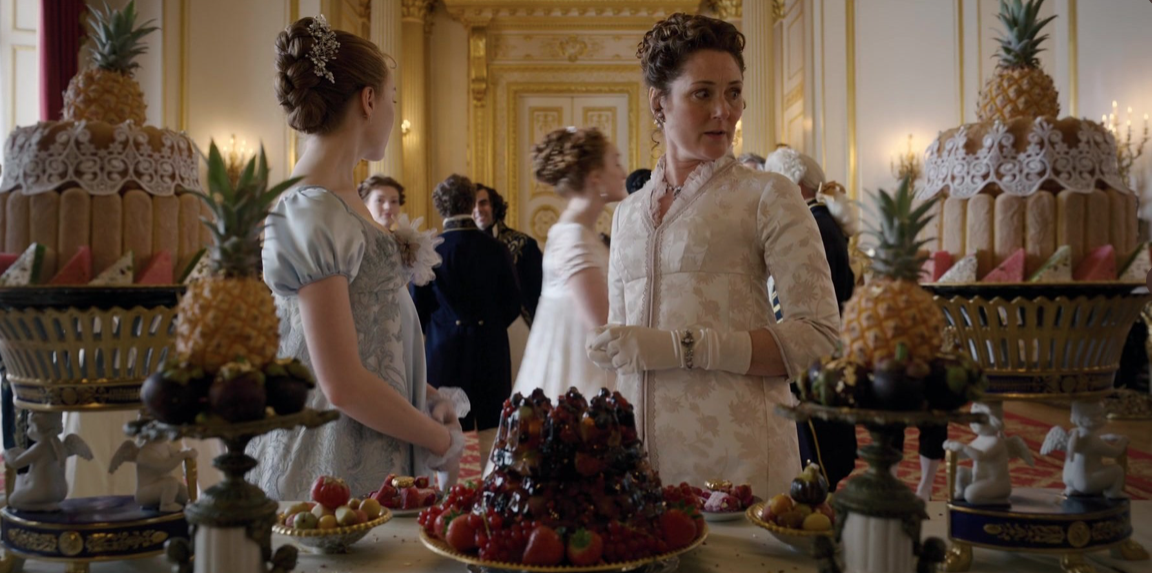 Picture shows: Lady Violet Bridgerton (Ruth Gemmell) and Daphne Bridgerton (Phoebe Dynevor) in front of a table of desserts that includes a jelly, two big cakes, and four pineapples.