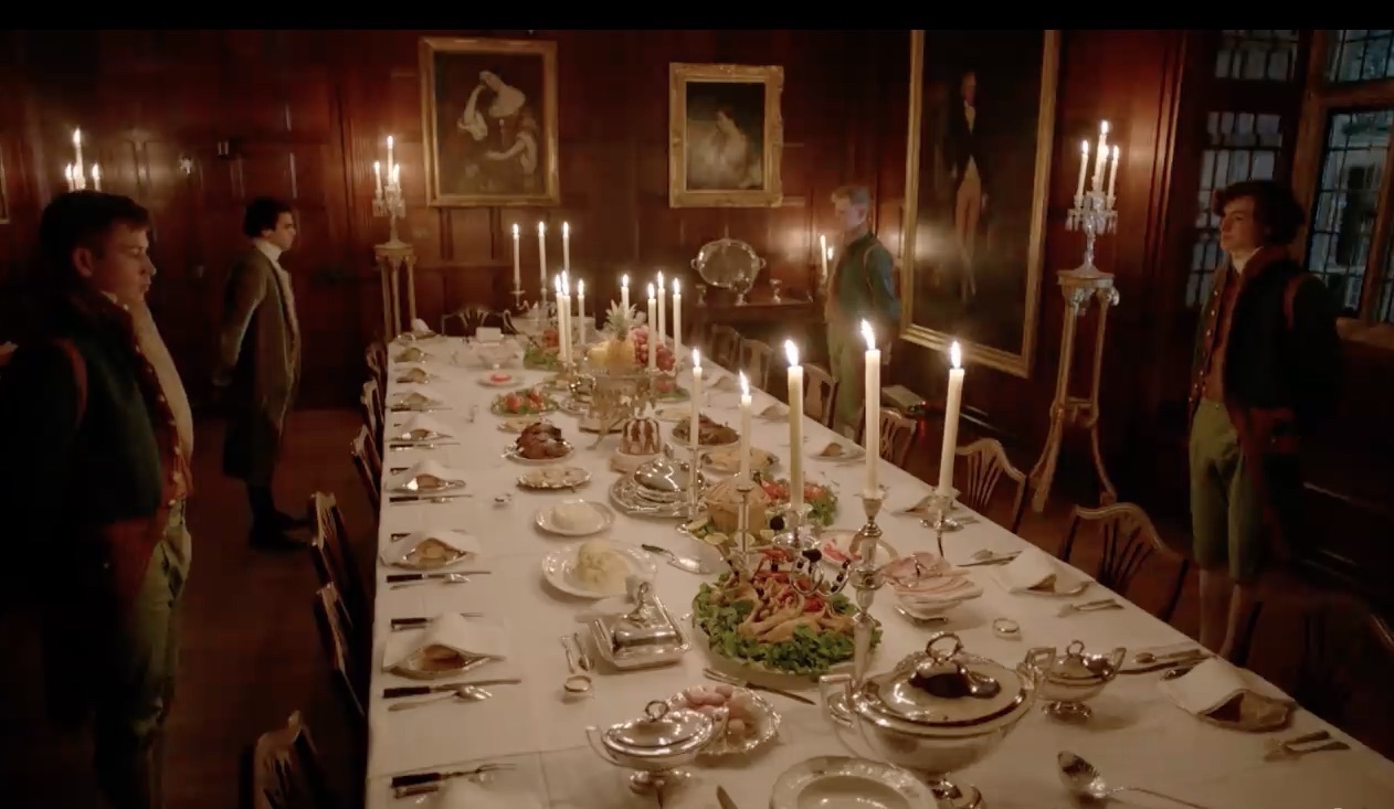 Picture shows: Footmen stand by to welcome guests to dinner. Food is arranged on the large table in a dining room that is lit by candlelight with portraits hanging on the wall.