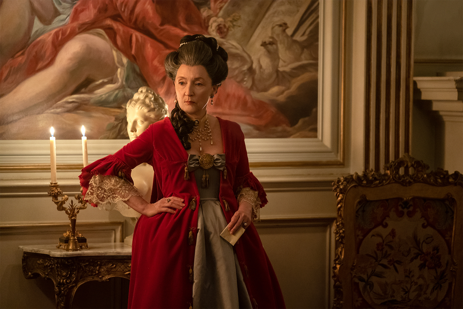 Picture shows: Genevieve de Merteuil (Lesley Manville) dressed in a red gown and holding a letter, stands in front of a painting in her opulent house.