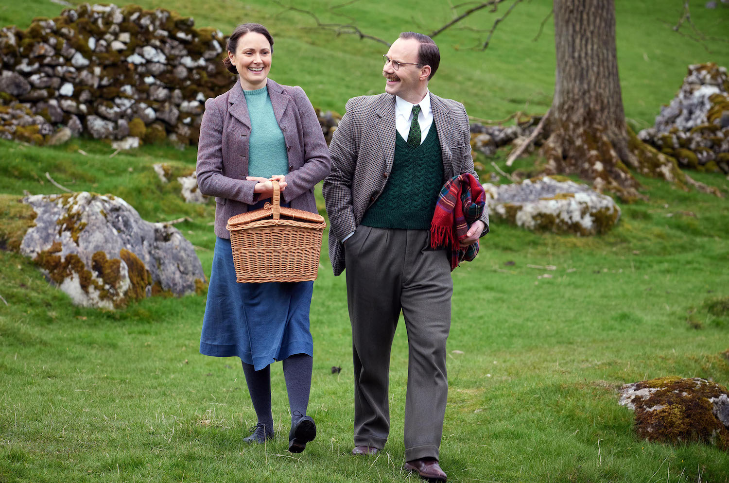 Picture shows: Mrs. Hall (Anna Madeley) and Gerald Hammon (Will Thorp) walk across a green field, with drystone walls, rocks, and a tree in the background. Mrs. Hall carries a picnic basket and Geral carries a folded rug. They both look very happy.