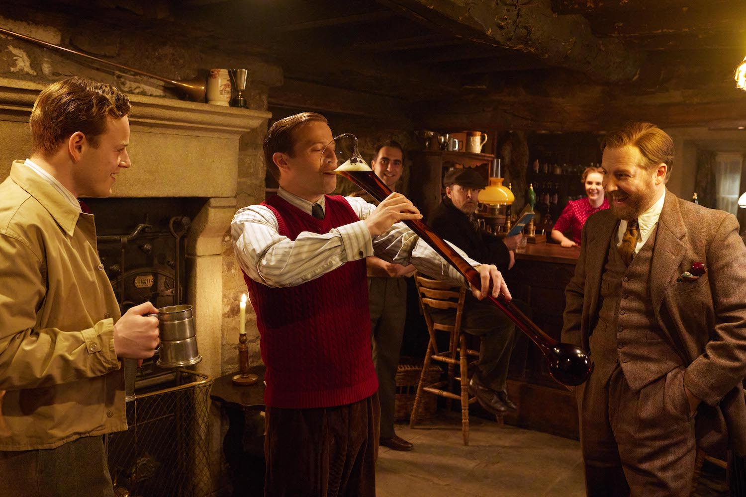 Picture shows: Tristan (Callum Woodhouse) and Siegfried (Samuel West) stand on either side of James (Nicholas Ralph) as he chugs a yard of ale in the pub. They are laughing and encouraging him.