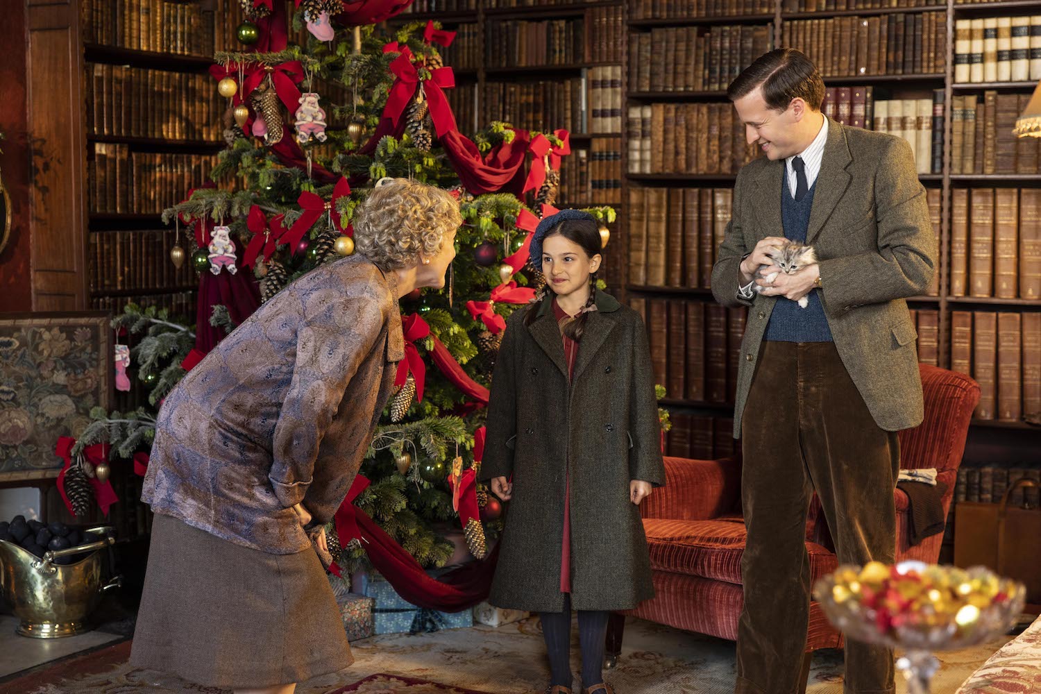 In an opulent book-lined room, Mrs. Pumphrey (Patricia Hodge) talks to Eva (Ella Bernstein) while James holds a small gray kitten. The room is decorated for Christmas