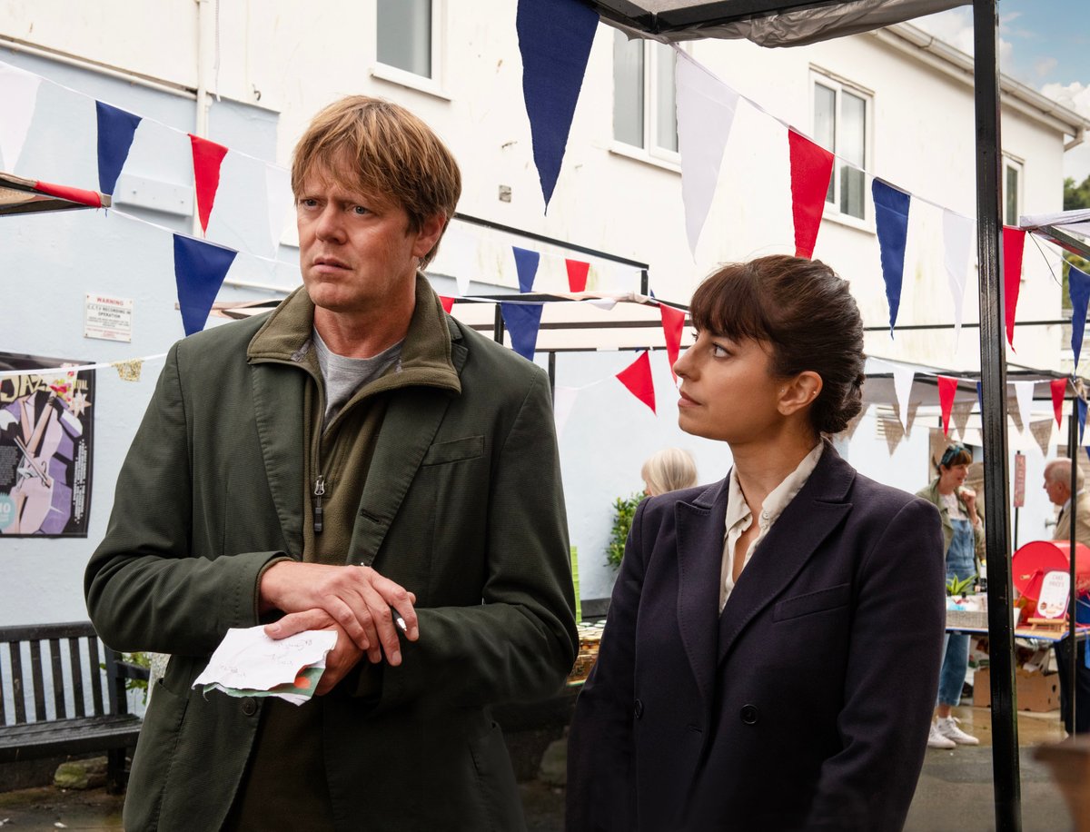 Picture shows: DI Humphrey Goodman (Kris Marshall) and DS Esther Williams (Zahra Ahmadi) stand in a street with white-walled houses nearby and red and blue bunting overhead. Humphrey looks thoughtful. He is holding a bundle of pieces of paper, and Esther is watching him think.