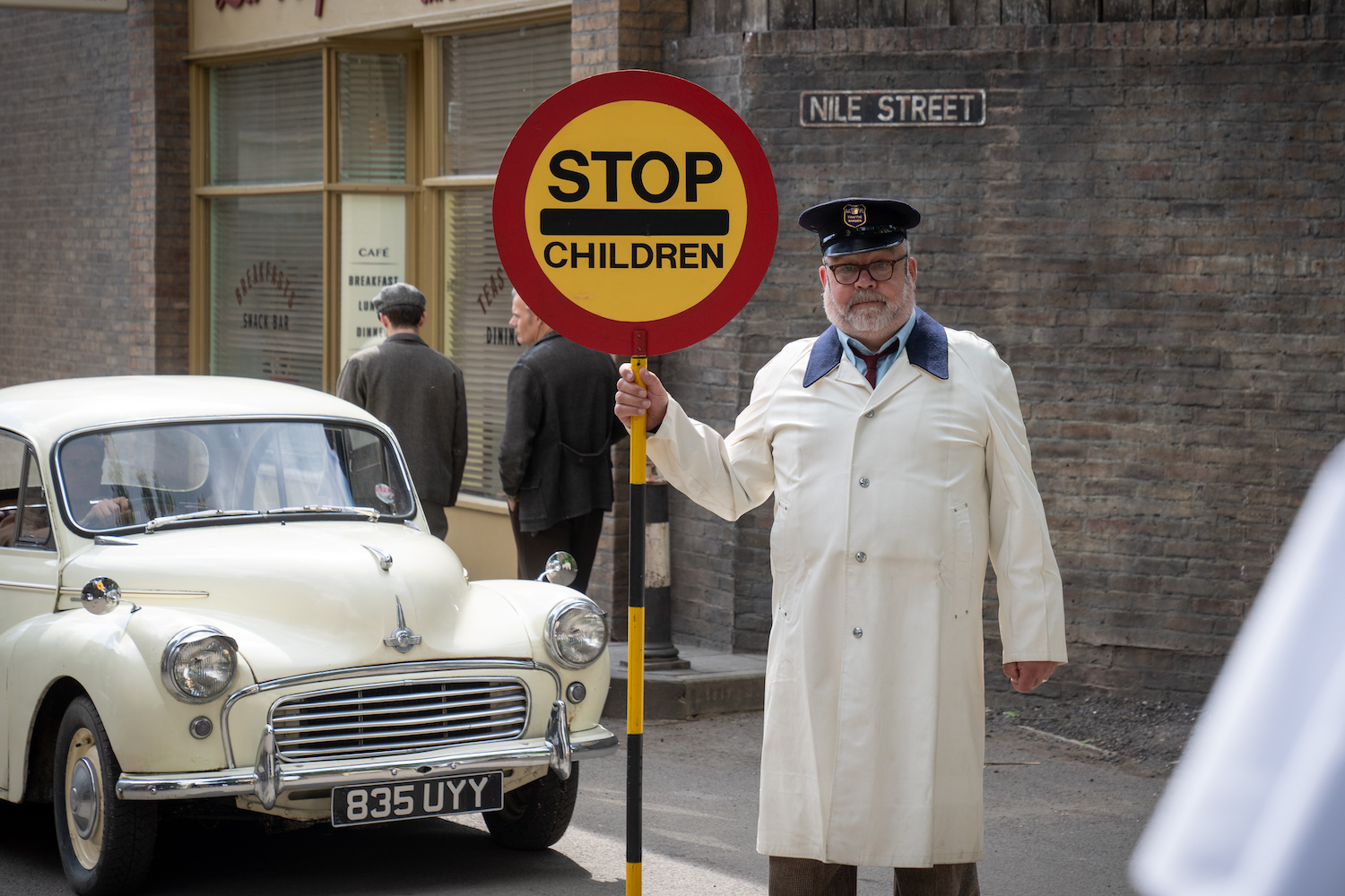 Picture shows: Fred Buckle (Cliff Parisi) in his new job. He's wearing a white coat and a black cap and holding a sign that reads "STOP-CHILDREN." Because the sign is round, it looks like a lollipop, hence the name "lollipop man."