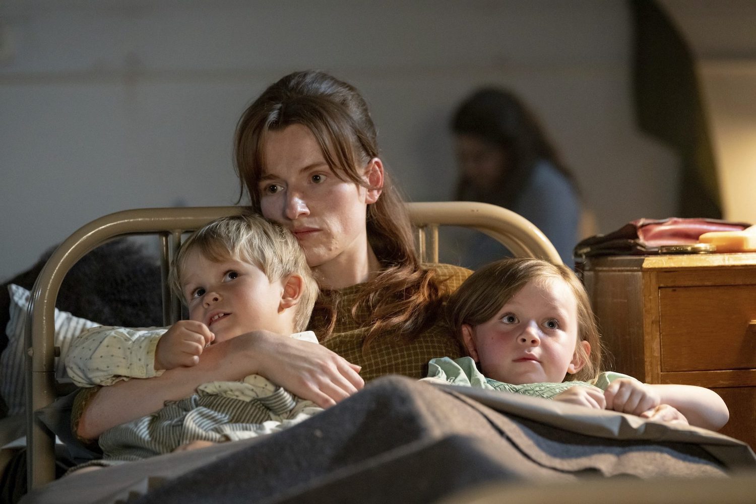 Picture shows: Sandy (Rose Riley) with her children at the shelter. They are cuddled up in an old-fashioned, narrow bed with Peter (Fred Jones) on Sandy's right and Ann Marie (Maisie Squibb) on her left.