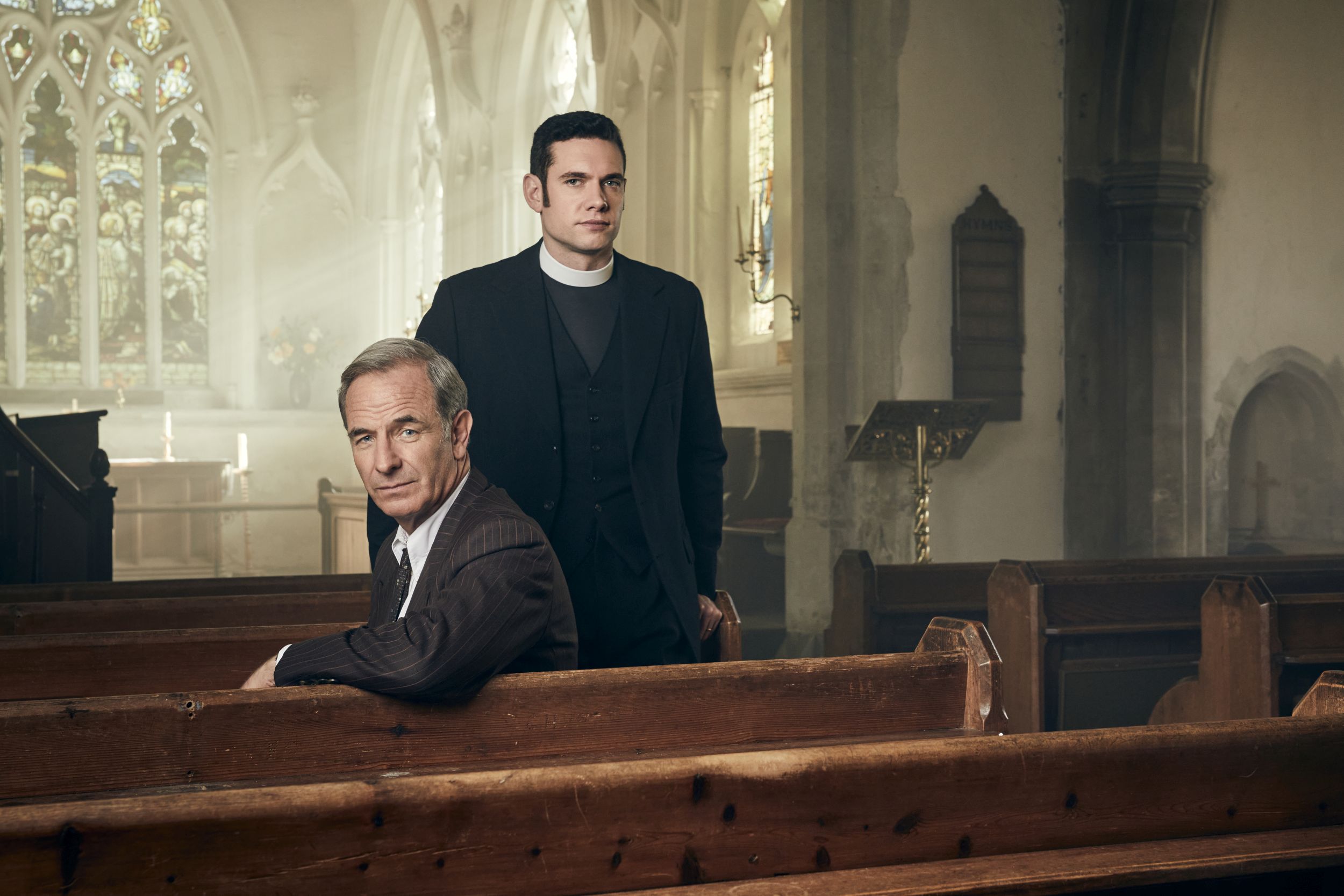 Tom Brittney as Will Davenport and Robson Green as Geordie Keating sitting in church pews in 'Grantchester' Season 8's Key Art 