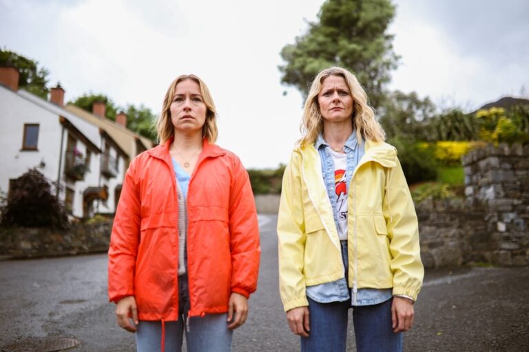 Sarah Goldberg and Susan Stanley as Sare and Suze in 'SisterS.' They are standing side-by-side in the middle of a street of houses.