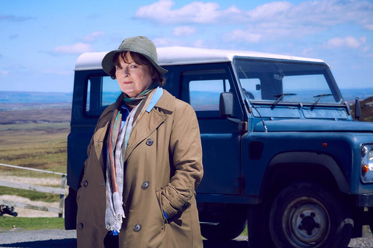 Vera stands in her classic hat and jacket in front of a car with green hills in the background.