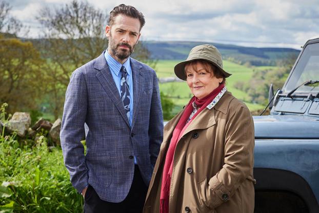 Vera and Joe stand together in front of a car with green hills behind them