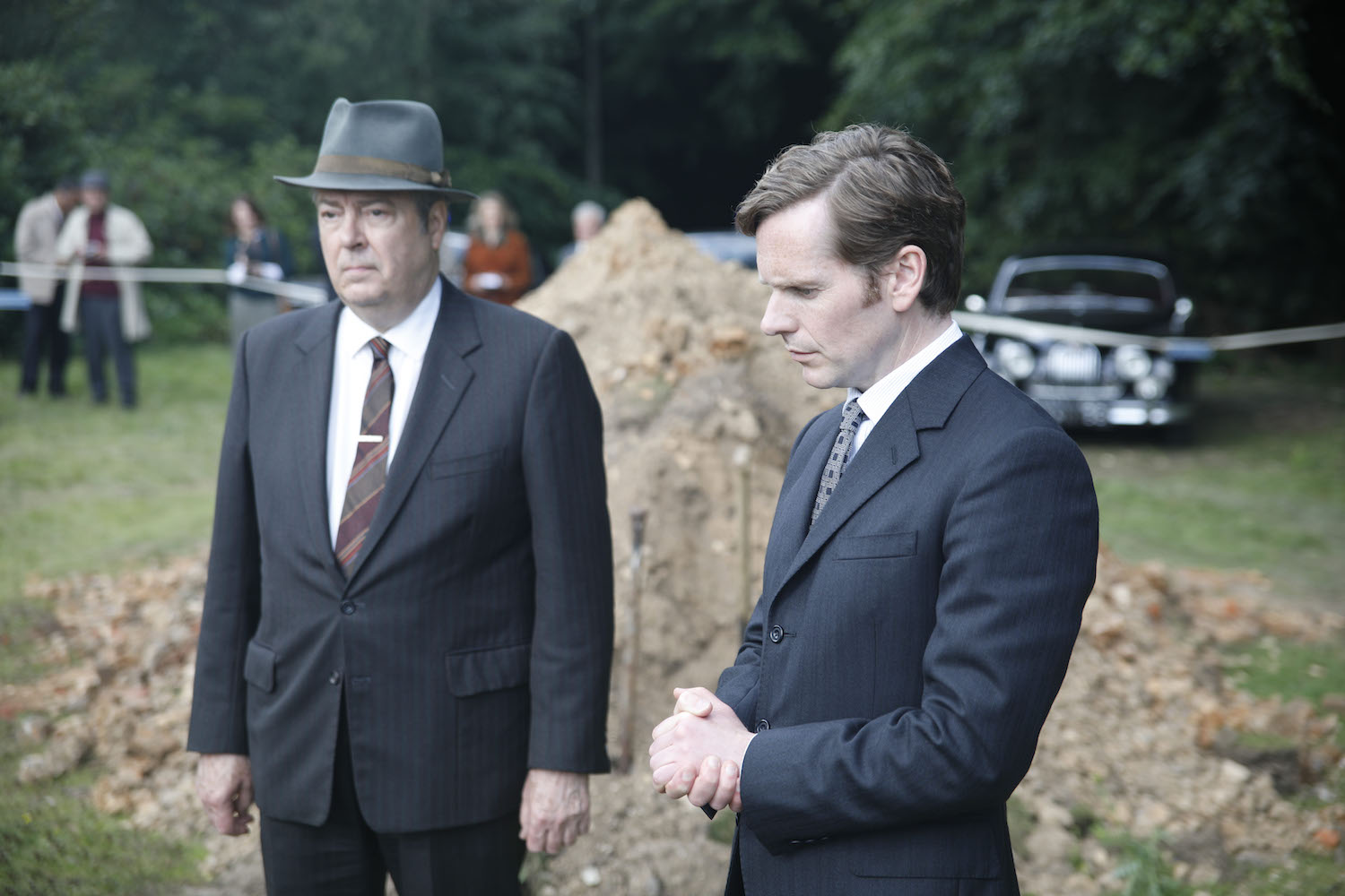 Picture shows: Fred Thursday (Roger Allam) and Morse (Sean Evans) at the excavation site in front of a large heap of dirt. In the background, journalists observe from behind a rope.