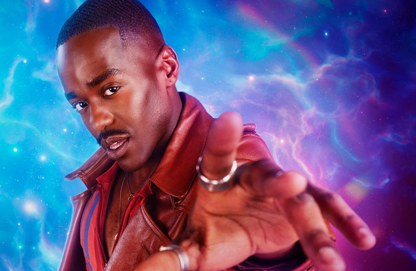A promo photo of Ncuti Gatwa, holding his hand out towards the camera. He is wearing a brown leather coat and a striped shirt, with silver rings on his hand. The background is a swirling blue and purple galaxy.