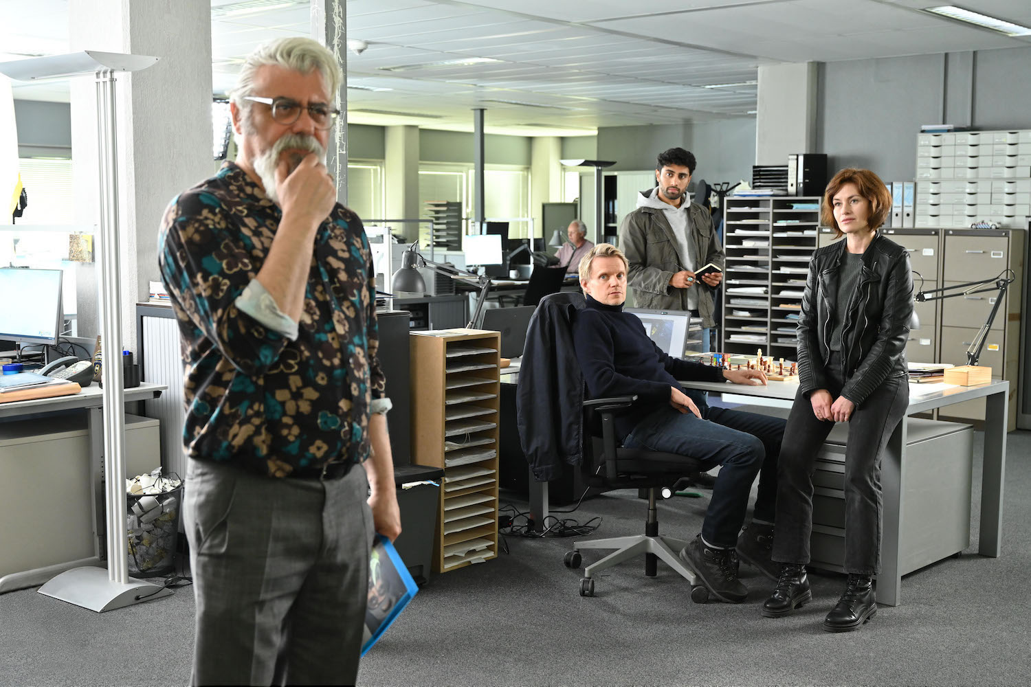 Picture shows: Pathologist Hendrik Davie (Darrel D'Silva) ponders the case while Piet Van der Valk (Marc Warren), seated at his desk, Eddie Suleman (Azan Ahmed), and Lucienne Hassell (Mamie McCoy) await his findings.