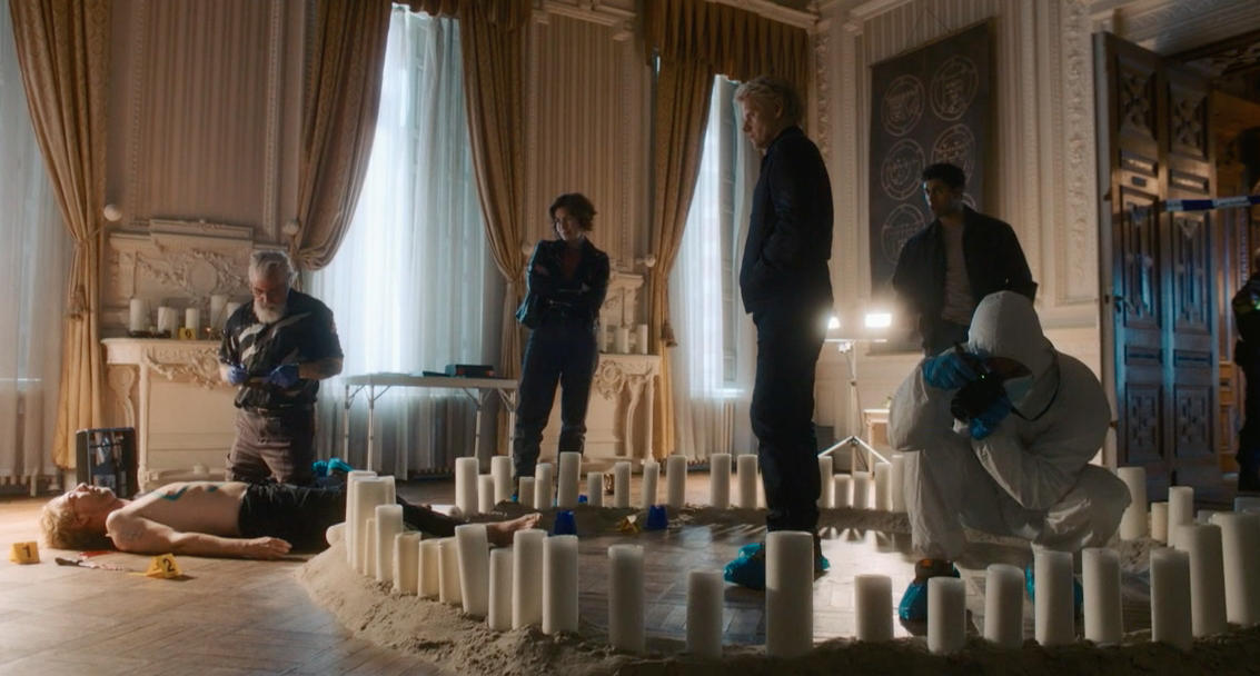 Picture shows: Van der Valk (Marc Warren) and his team investigate the murder of Nik Delacorte (Joris Smit). Hendrik Davie (Darrell D’Silva) examines the body which is in an opulently-furnished room with elaborate plasterwork and heavy curtains. A crime scene photographer kneels in a circle of pillar candles standing in sand. Lucienne Hassell (Mamie McCoy) and Eddie Suleman (Azan Ahmed) observe.