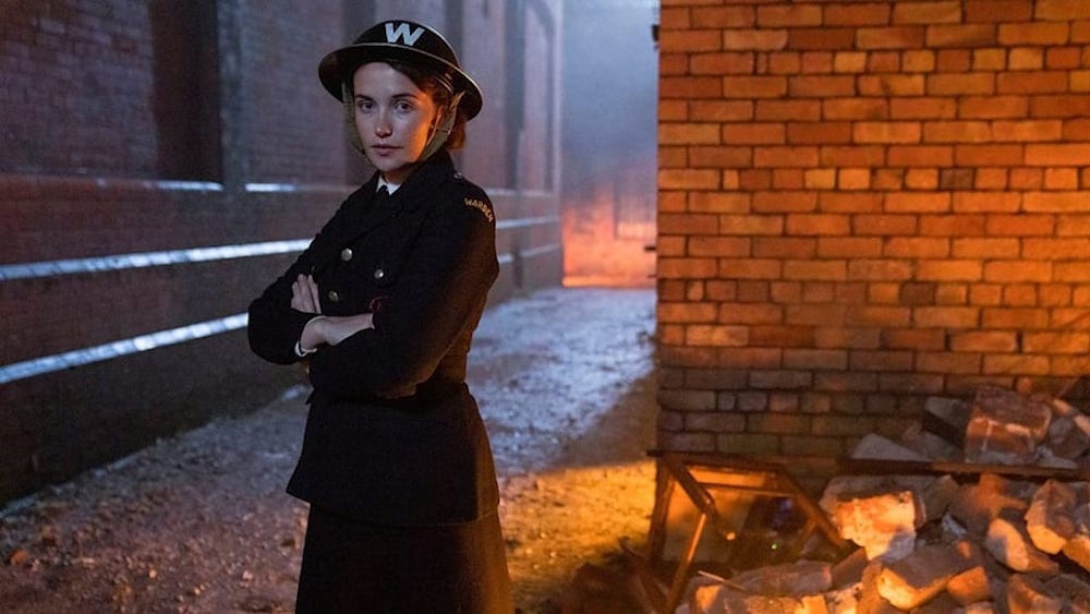 Picture shows: Julia Brown as Lois. In her dark ambulance driver uniform, she stands arms folded in front of a brick wall among rubble. There are buildings on fire in the background
