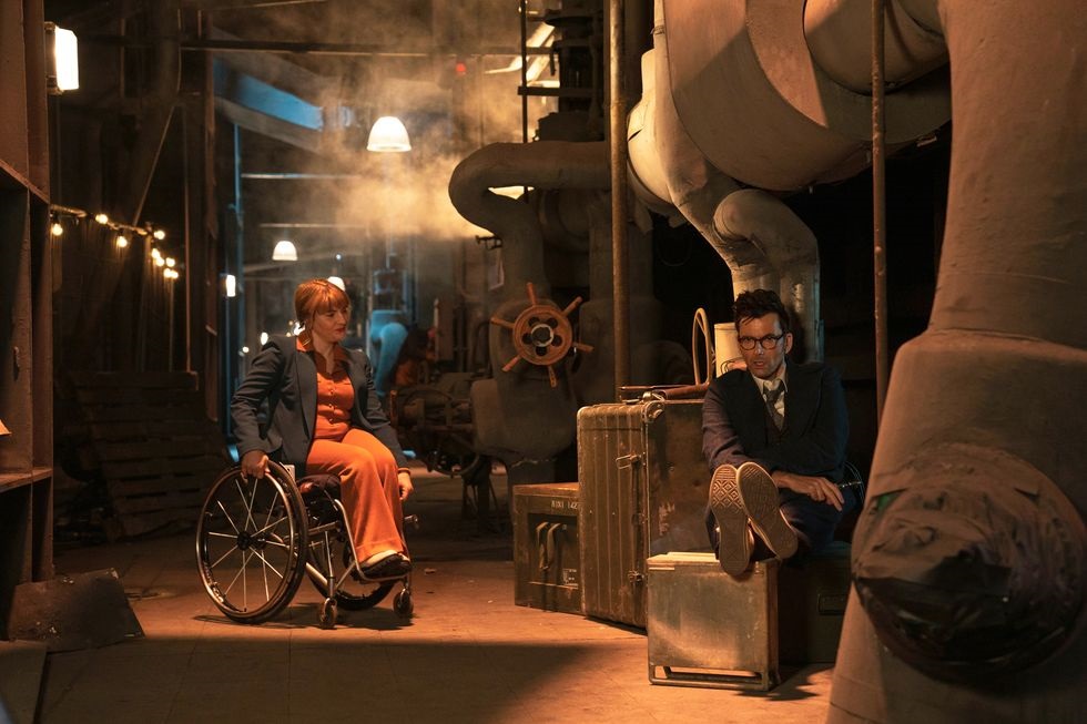 Shirley (Ruth Madeley) sits in her wheelchair with her legs crossed, speaking with the Doctor (David Tennant) who is seated and holding a tablet.