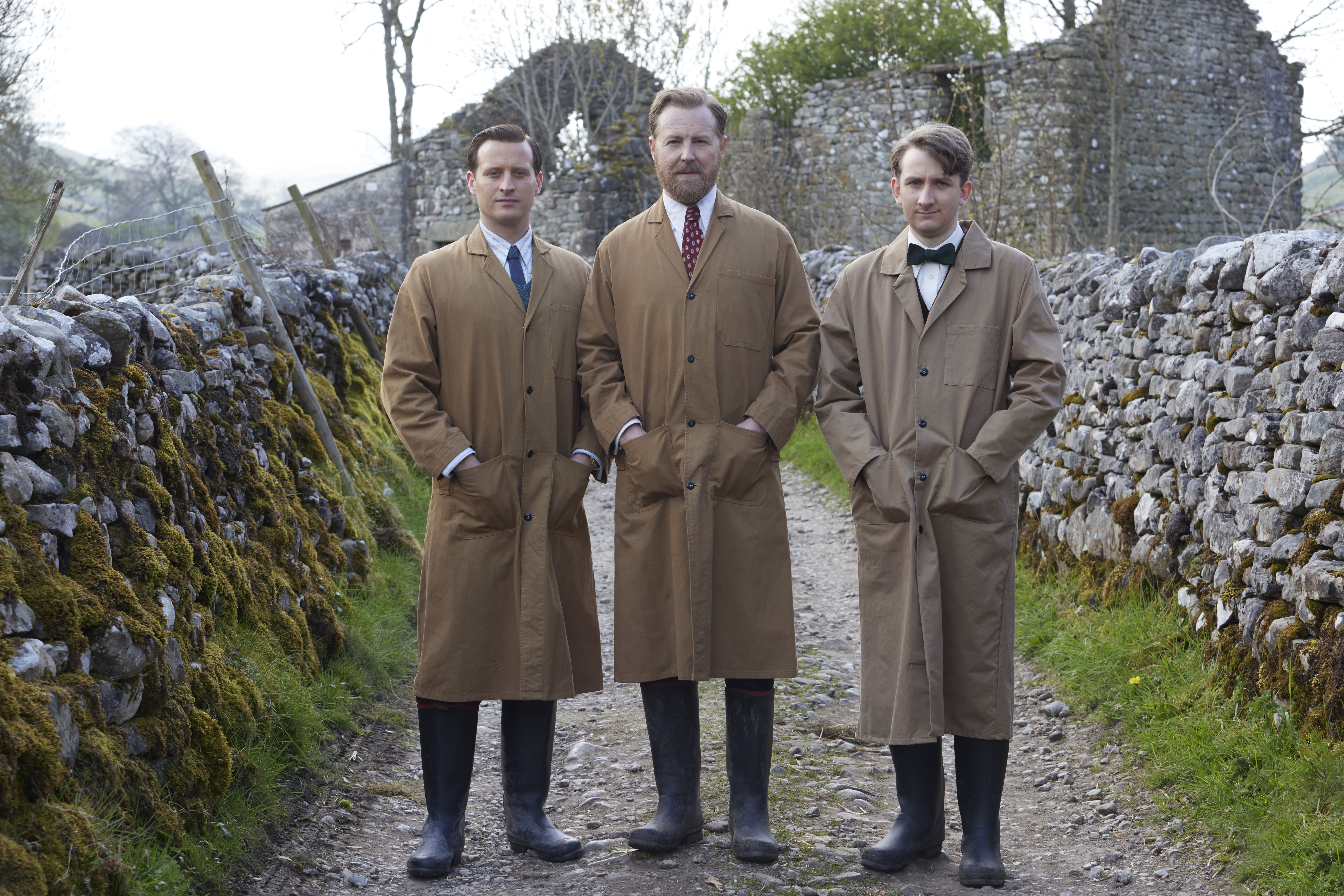 Picture shows: Dressed to clean, James Herriot (Nicholas Ralph), Siegfried Farnon (Samuel West), and Richard Carmody (James Anthony-Rose) stand in a stony lane edged with drystone walls. They are wearing heavy-coats for messy work.
