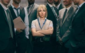 Picture shows: A small blond woman, arms crossed, stands in an elevator full of men. She barely reaches their shoulders.
