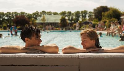 Picture shows: At an outdoor swimming pool Tom (Harry Styles) and Marion (Emma Corrin) smile at each other as they rest their elbows on the edge of the pool.