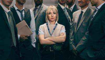 Picture shows: A small blond woman, arms crossed, stands in an elevator full of men. She barely reaches their shoulders.