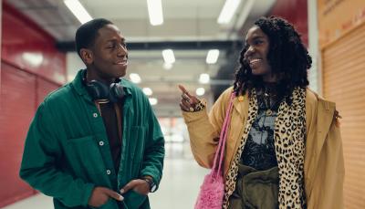 Dom (David Jonsson) and Yas (Vivian Oparah) smile at each other, talking animatedly, as they walk down the hallway of an indoor market