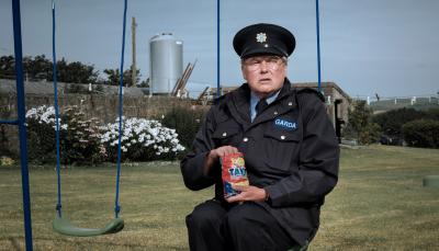 Picture shows: Sgt. P.J. Collins (Conleth Hill), wearing his Garda uniform, sits on a child's swing eating chips from a bag. He looks disconsolate.