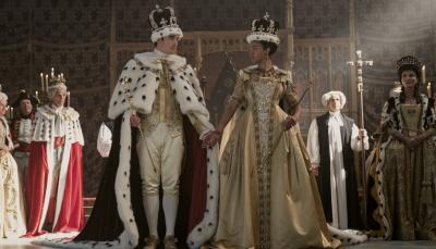 Corey Mylchreest as Young King George and India Amarteifio as Young Queen Charlotte take the throne together for the first time as Michelle Fairley as Princess Augusta looks on in Queen Charlotte: A Bridgerton Story