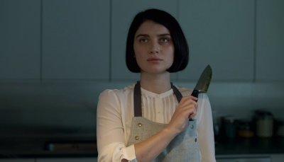 Eve Hewson as Adele Ferguson wears an apron and holds up a knife defensively in a darkened room