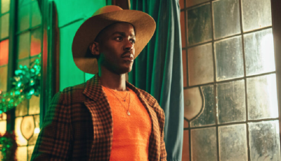 Ncuti Gatwa wears an orange sweater, brown suit, and cowboy hat. He stands indoors next to a foggy window.