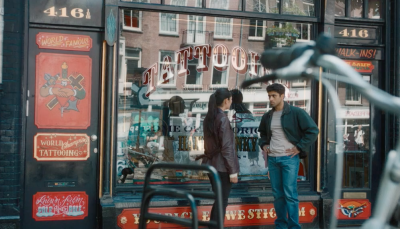 Picture shows: Citra Li (Django Chan-Reeves) listens as Eddie Suleman (Azan Ahmed) shares his bad experience with tattooists. They are outside a tattoo shop, with parked motorcycles in the foreground. The shop is bright and eye-catching.