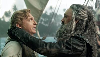 On a ship in a harbor, Stede (Rhys Darby) looks on fearfully as Blackbeard (Taika Waititi) holds him by the neck