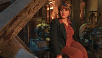 Ruth Madeley as Shirley, a white woman in her 30s wearing a red jumpsuit and dark blazer seated in a wheelchair among industrial debris