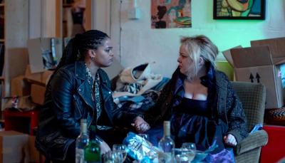 Eddie (Lydia West) looks concerned at Maggie (Nicola Coughlan), who looks unkempt. They are sitting in the middle of a messy apartment.