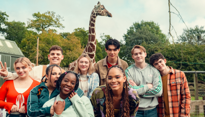 The cast of Heartstopper, including the characters Charlie, Nick, Tao, Elle, Darcy, Tara, and Isaac, pose for a photo together at a zoo with a giraffe behind them. All are smiling.