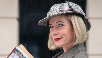 Lucy Worsley in a deerstalker hat and book in hand in front of the 221B Baker Street door in 'Lucy Worsley's Holmes vs. Doyle.'