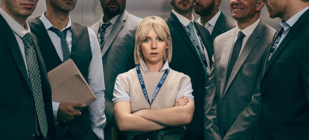 Picture shows: A small blond woman, arms crossed, stands in an elevator full of men. She barely reaches their shoulders.