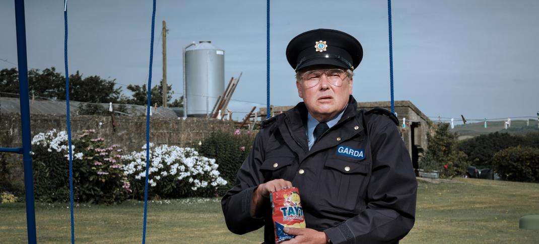 Picture shows: Sgt. P.J. Collins (Conleth Hill), wearing his Garda uniform, sits on a child's swing eating chips from a bag. He looks disconsolate.