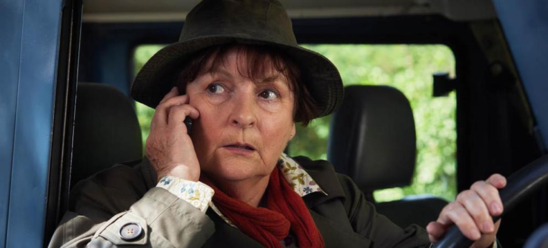 Brenda Blethyn as DCI Vera Stanhope. She is sitting in the drivers' seat of a car with one hand on the steering wheel and another holding a phone to her ear.