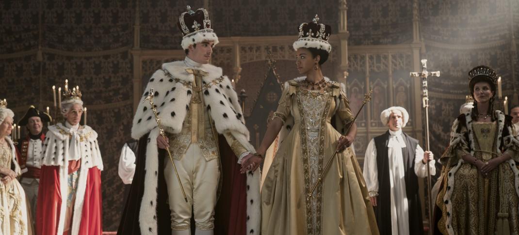 Corey Mylchreest as Young King George and India Amarteifio as Young Queen Charlotte take the throne together for the first time as Michelle Fairley as Princess Augusta looks on in Queen Charlotte: A Bridgerton Story