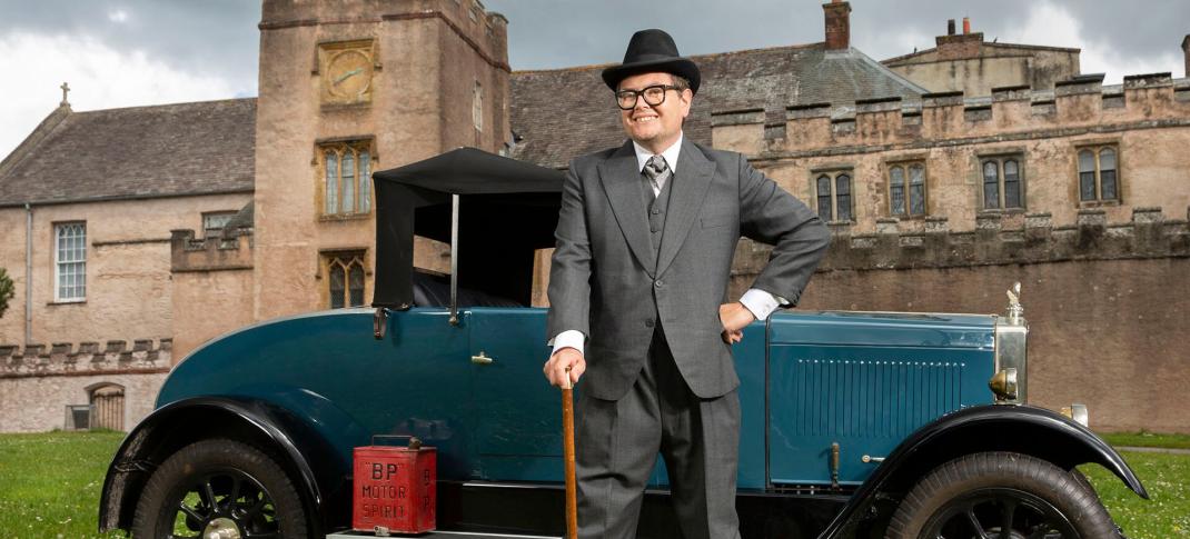Alan Carr stands in front of a 1920s car and estate in Alan Carr's Adventures with Agatha Christie