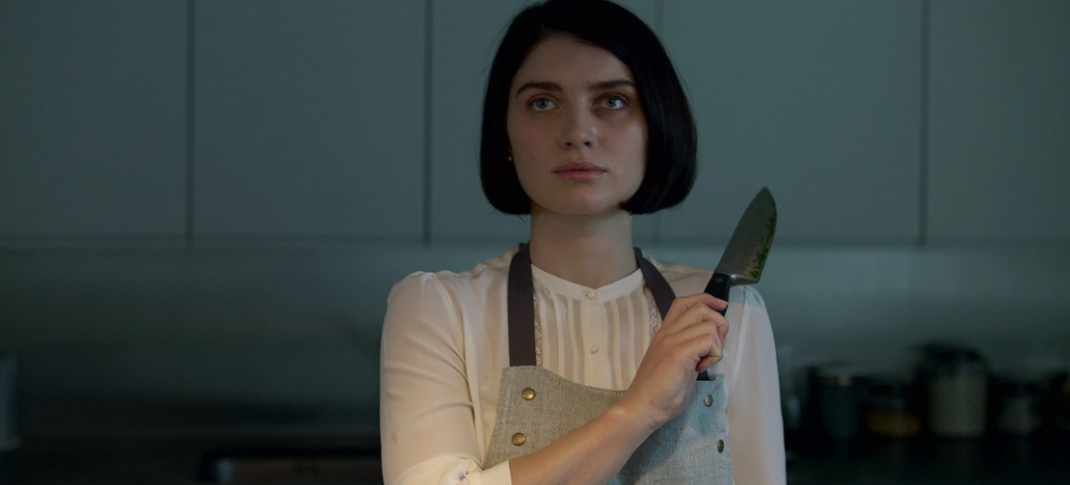 Eve Hewson as Adele Ferguson wears an apron and holds up a knife defensively in a darkened room