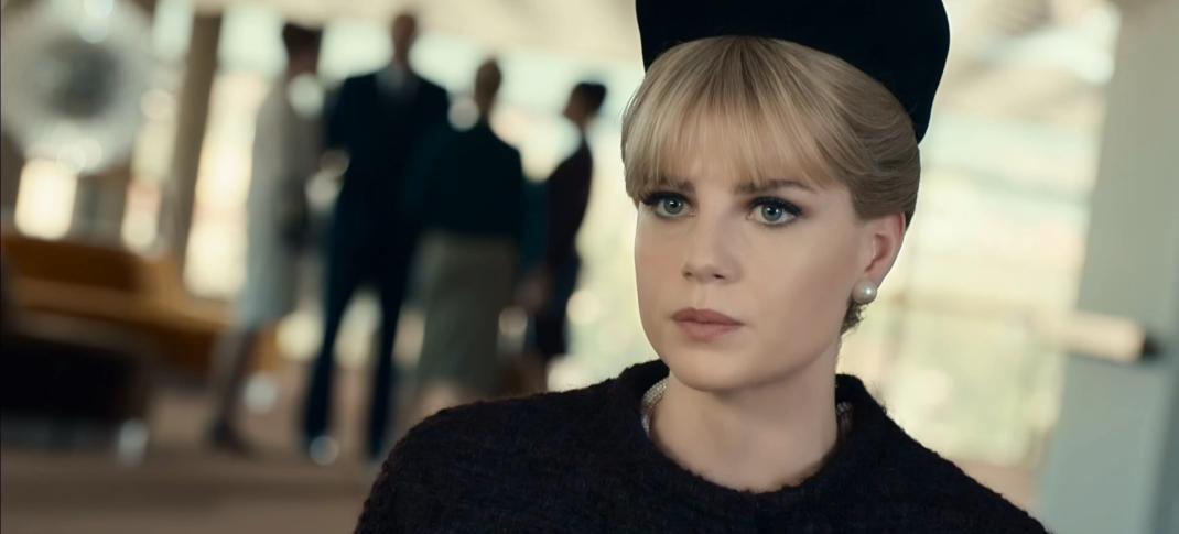 A closeup of Lucy Boynton in a 1960s style black hat and black coat, with a group of people also dressed in dark clothes out of focus in the background.