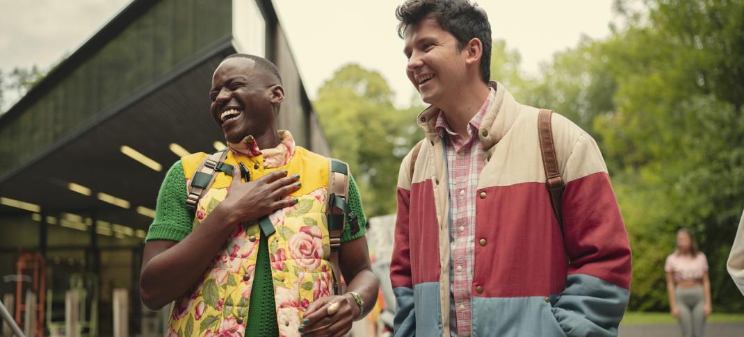 Eric (Ncuti Gatwa) and Otis (Asa Butterfield) stand together outside a school building laughing and smiling