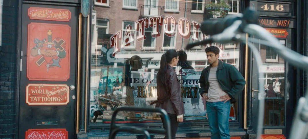 Picture shows: Citra Li (Django Chan-Reeves) listens as Eddie Suleman (Azan Ahmed) shares his bad experience with tattooists. They are outside a tattoo shop, with parked motorcycles in the foreground. The shop is bright and eye-catching.