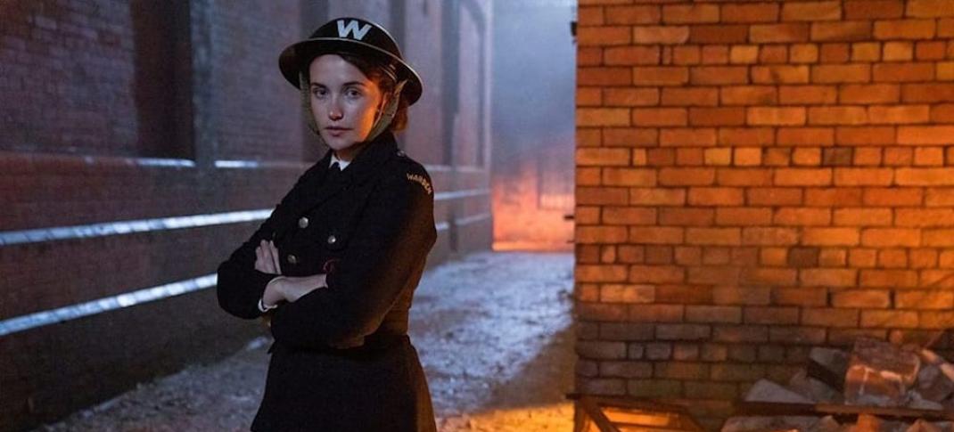 Picture shows: Julia Brown as Lois. In her dark ambulance driver uniform, she stands arms folded in front of a brick wall among rubble. There are buildings on fire in the background