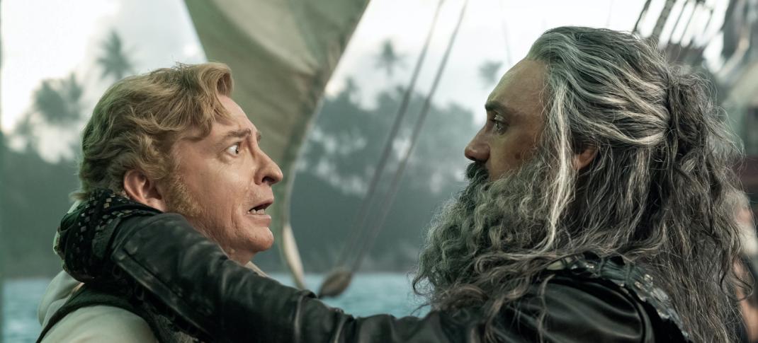 On a ship in a harbor, Stede (Rhys Darby) looks on fearfully as Blackbeard (Taika Waititi) holds him by the neck