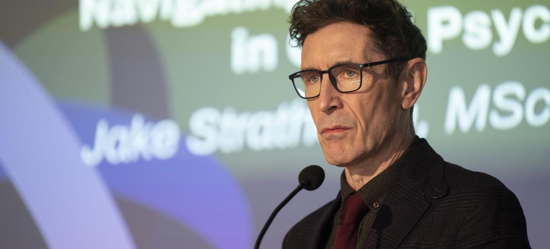 Paul McGann as Jake Strathearn at a lectern in 'Annika' Season 2