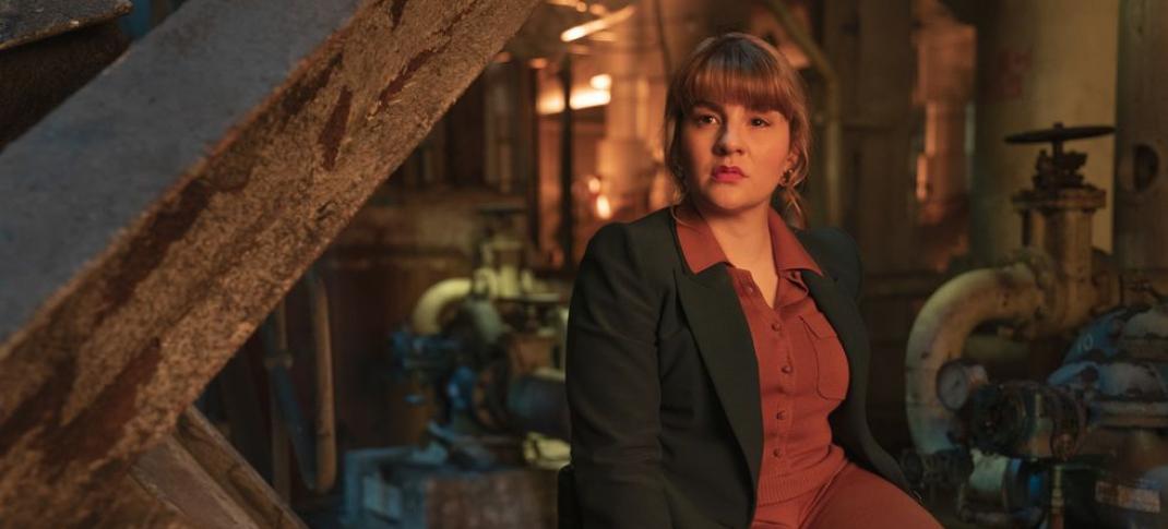 Ruth Madeley as Shirley, a white woman in her 30s wearing a red jumpsuit and dark blazer seated in a wheelchair among industrial debris