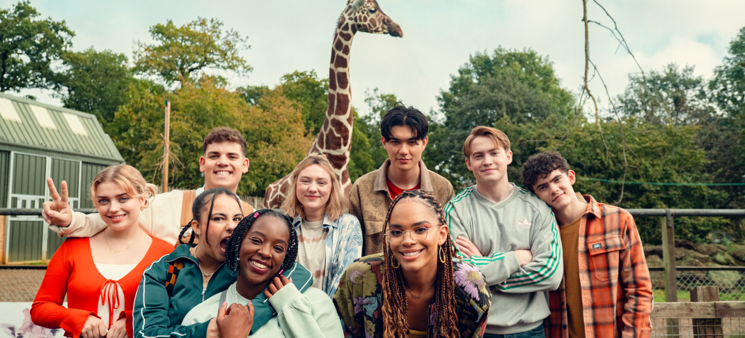 The cast of Heartstopper, including the characters Charlie, Nick, Tao, Elle, Darcy, Tara, and Isaac, pose for a photo together at a zoo with a giraffe behind them. All are smiling.