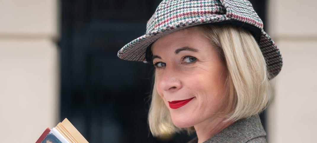 Lucy Worsley in a deerstalker hat and book in hand in front of the 221B Baker Street door in 'Lucy Worsley's Holmes vs. Doyle.'